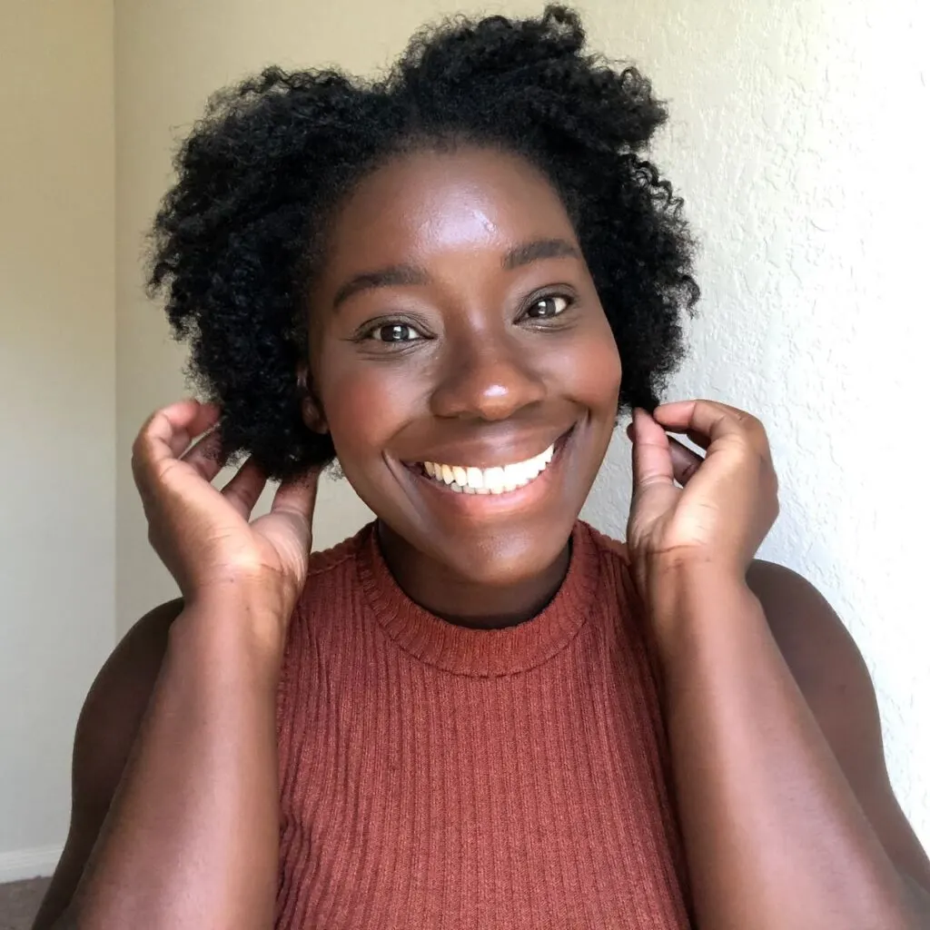 black woman smiling into the camera while holding her natural 4c afro in hand