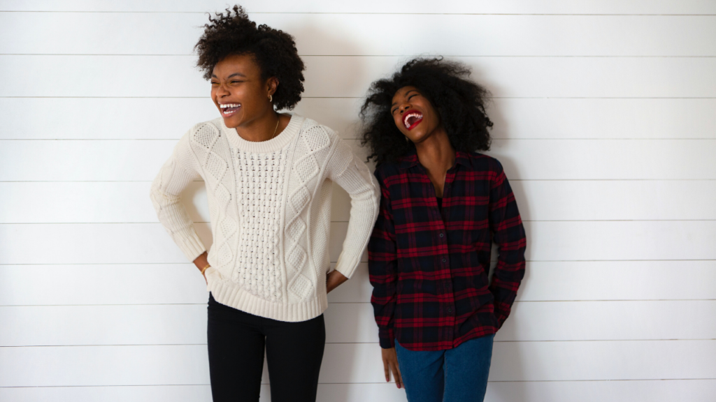 two young black women standing next to each other in front of white wall laughing