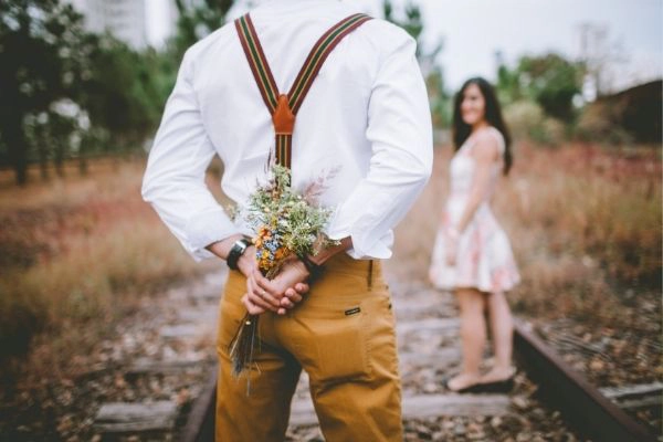 young man standing with his back to the camera while holding a flower behind his back and looking at young woman