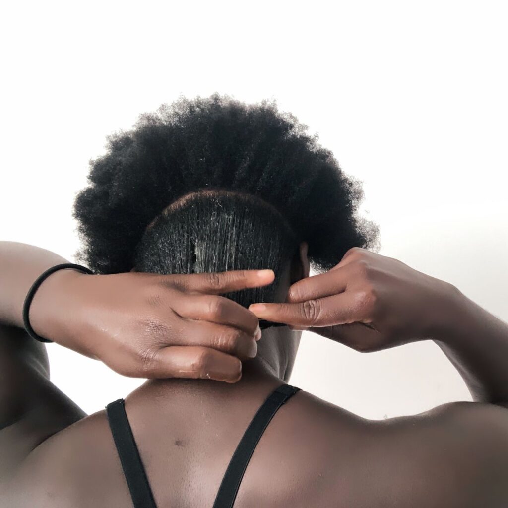 black woman applying edge control to natural 4c hair section by section using a wide tooth comb and fingers. 