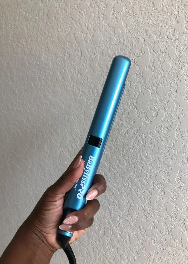 Black hand holding the Babyliss Pro Nano Titanium Flat Iron in front of white wall