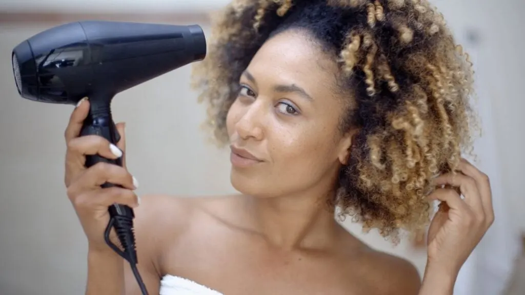 black woman with curly blonde hair holding a black blow dryer