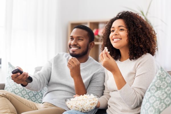 black couple sitting on the couch eating popcorn with tv remote in hand